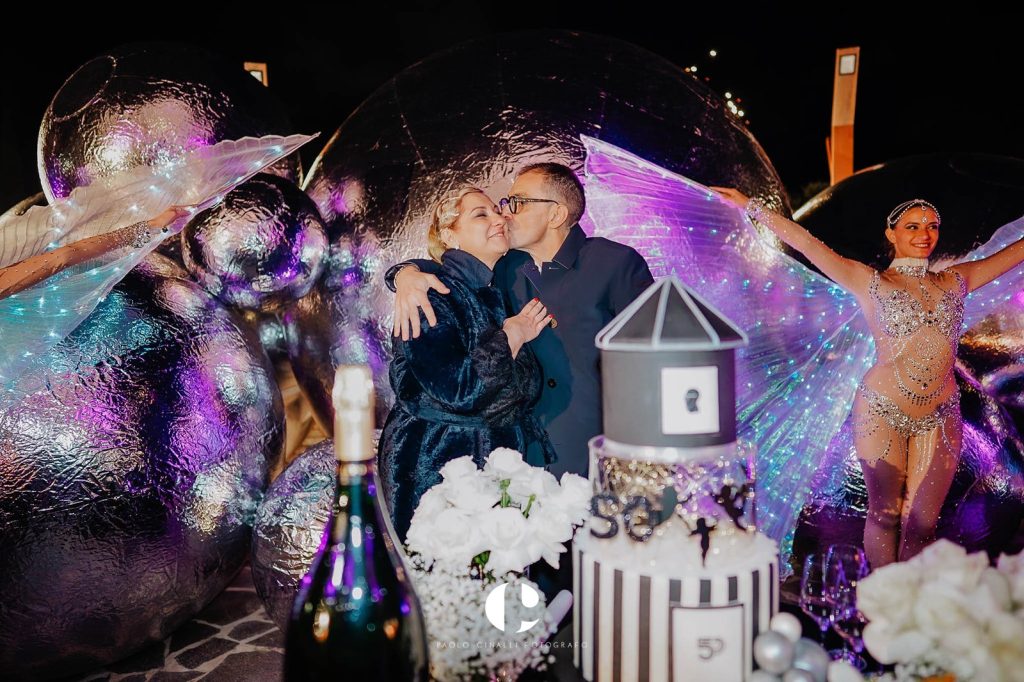 Un bacio accanto alla torta, tra le sfere specchiate e i performer con le ali di luce